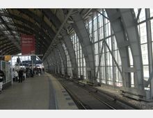 Bahnsteig einer überdachten Bahnhofshalle mit großen Stahlträgern und Glaswänden, durch die Tageslicht einfällt. Im Hintergrund sind Fahrpläne, Hinweisschilder und wartende Passagiere sichtbar.