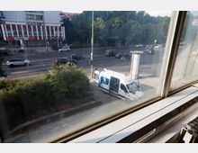 Blick aus einem Fenster auf eine städtische Straßenkreuzung mit fließendem Verkehr, einem geparkten weißen Transporter mit geöffneter Seitentür und einem Gehweg mit Büschen im Vordergrund. Im Hintergrund ist ein mehrstöckiges Gebäude mit rot-weißer Fassade und Bäume entlang der Straße zu sehen.