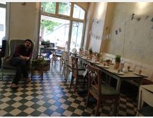 Ein gemütliches Café-Interieur mit kariertem Boden, langen Gemeinschaftstischen, die mit Tellern, Gläsern und kleinen Kräutertöpfen dekoriert sind, sowie einem großen Sessel im Vordergrund. Im Hintergrund sind Türen zu einem Außenbereich geöffnet, durch die Grünflächen sichtbar sind.