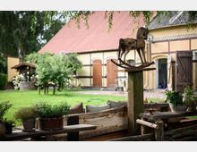 Hof mit historischem Fachwerkgebäude im Hintergrund, dazu ein Vorgarten mit Rasen, Zierpflanzen und einem rustikalen Sitzbereich aus Holz. Im Vordergrund steht eine dekorative Holzskulptur in Form eines Schaukelpferds auf einem Pfosten.