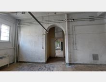 Ein leerer, verlassener Raum mit abgenutzten Wänden, sichtbaren Rohren und einfacher Beleuchtung. Im Vordergrund ist ein Türdurchgang mit einem bogenförmigen Abschluss erkennbar, der zu weiteren Räumen mit ähnlichem Zustand führt.