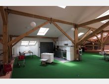 Wohnraum in einem Dachgeschoss mit sichtbaren Holzbalken, grünem Teppichboden und mehreren Oberlichtern. Im Vordergrund befindet sich eine offene Küche mit weißen Schränken, daneben ein Essbereich und ein Arbeitsplatz.