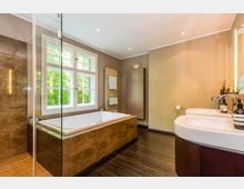 Modernes Badezimmer mit großer Badewanne in braunem Marmordesign, bodentiefer Glasdusche, Doppelwaschbecken und einem weißen Fenster mit Blick ins Grüne. Der Holzboden und die warmen Wandfarben sorgen für eine einladende Atmosphäre.