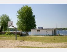 Eine Sandfläche mit einem Basketballkorb im Vordergrund, daneben ein weißes Gebäude mit der Aufschrift "BOOTE". Im Hintergrund sind Bäume, ein See und einige Segelboote zu sehen.