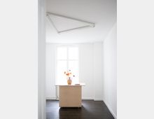 Minimalistisch eingerichteter Raum mit weißen Wänden und dunklem Holzboden. Im Vordergrund steht ein schlichter Holztisch mit einer Keramikvase mit orangefarbenen Blumen, im Hintergrund ein großes, bogenförmiges Fenster.