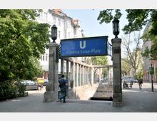 Eingang zur U-Bahn-Station Viktoria-Luise-Platz in Berlin, mit einem blauen Schild und steinernen Säulen. Im Hintergrund sind Bäume, Wohnhäuser und geparkte Fahrräder zu sehen.