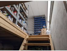 Holztreppe, die zu einem kleinen Lagerraum mit Regalen führt. Die Regale enthalten verschiedene Behälter, Dosen und Vorratsboxen, darunter Werkstattbedarf, während auf der rechten Seite eine Betonwand und ein schräg einfallendes Oberlicht zu sehen sind.