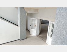 Treppenhausbereich eines Gebäudes mit hell gefliesten Böden, weißen Wänden und einer offenen Tür zu einem angrenzenden Raum mit Betonboden. Ein Treppengeländer und eine verzierte tragende Wand mit grauer Struktur sind sichtbar.