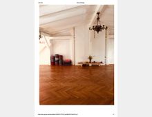 Ein heller Raum mit weiß gestrichenen Holzbalken an der Decke, einem Boden in Fischgrätparkett, einem Kronleuchter und einer schlichten Dekoration. Links stehen gestapelte Yogamatten und Kissen, rechts befindet sich ein niedriger Holztisch mit Pflanzen und Kerzen.