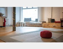 Heller Raum mit Holzfußboden und großen Teppichen, in der Mitte ein rotes Sitzkissen. Im Hintergrund eine niedrige Sitzbank mit Kissen vor einem großen Fenster, weiße Ziegelwand und ein Heizkörper.