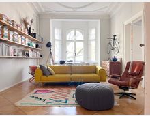 Wohnzimmer im Altbau mit hohen Decken und Stuckverzierungen, gelbem Sofa, rotem Ledersessel und modernem Dekor. An einer Wand befinden sich Holzregale mit Büchern, an einer anderen ein angebrachter Fahrradhalter; große weiße Fenster im Hintergrund lassen viel Tageslicht einfallen.