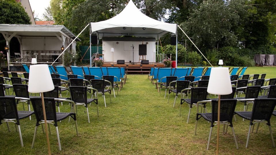 Open-Air-Veranstaltungsbereich auf einer Rasenfläche mit Reihen von Stühlen, die auf eine kleine Bühne unter einem weißen Pavillonzelt ausgerichtet sind. Hinter der Bühne stehen Bäume und Grünflächen, daneben ein überdachter Holzpavillon und ein Gebäude am Rand.
