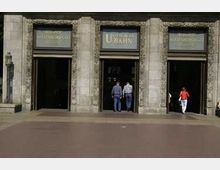 Eingang zu einer U-Bahn-Station in einem historischen Gebäude mit verzierten Steinsäulen und drei Türen, von denen zwei Personen betreten oder verlassen. Oben befindet sich ein Schild mit der Aufschrift "U-Bahn".
