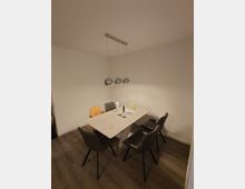 Essbereich mit rechteckigem Esstisch aus hellem Material, umgeben von vier gepolsterten, schwarzen Stühlen und einem hellbraunen Kinderstuhl. Über dem Tisch hängen drei silberne, kugelförmige Pendelleuchten, Boden aus dunklem Holzimitat, Wände schlicht weiß.