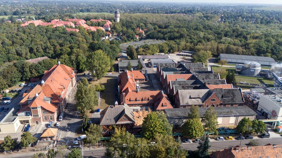 Luftaufnahme eines weitläufigen Industrie- und Fabrikgeländes mit mehreren Backsteinhallen und roten Ziegeldächern, umgeben von vielen Bäumen. Rechts sind technische Anlagen mit Rohrleitungen und runden Tanks zu sehen, im Hintergrund Waldflächen und eine Siedlung mit einem markanten Turm.
