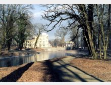 Ein gefrorener Teich inmitten eines Parks mit kahlen Bäumen im Winter; im Hintergrund ein helles, historisches Gebäude und ein pavillonähnliches Bauwerk. Der Boden ist mit Laub bedeckt, und ein schmaler Weg verläuft entlang des Teichs.