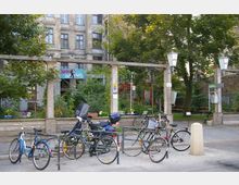 Mehrere Fahrräder und ein Motorroller sind vor einer kleinen urbanen Grünfläche abgestellt, die mit Bäumen, Pflanzen und Sitzmöglichkeiten gestaltet ist. Im Hintergrund ist eine Fassade eines älteren Gebäudes mit großen Fenstern zu sehen, und ein handgemaltes Schild mit der Aufschrift „Törtchen Backhaus“ hängt über dem Eingang.