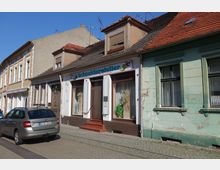 Reihenhäuser in einer Altstadt mit einem kleinen Schneiderei-Geschäft in der Mitte, gekennzeichnet durch ein Schild mit der Aufschrift „Schneideratelier“. Die Gebäude haben teils renovierte, teils abblätternde Fassaden, und ein geparktes Auto steht am Straßenrand.