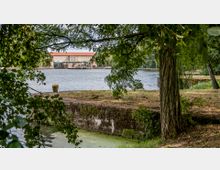Ein ruhiger Fluss mit einem Uferbereich, gesäumt von Bäumen und Grasflächen, im Vordergrund. Im Hintergrund ist ein großes Gebäude mit roten Ziegeldächern und großen Fenstern sichtbar.