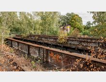 Verfallene Eisenbahnbrücke mit rostiger Struktur und Graffiti, umgeben von dichtem Laub und Bäumen; im Hintergrund ein altes Gebäude mit leuchtend gelben Fenstern.