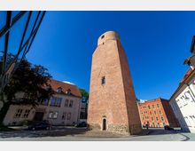 Backsteinturm in einem städtischen Umfeld mit blauem Himmel, umgeben von historischen Gebäuden mit roten Ziegeldächern und Fassaden aus Putz oder Backstein; der Turm steht auf einem Fundament aus Feldsteinen.