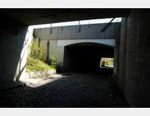 Unterführung mit gepflastertem Weg und Betonwänden, teils im Schatten. Im Hintergrund ist eine kleine Grünfläche mit Pflanzen und ein Schimmer von Tageslicht zu sehen.