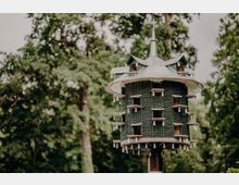 Zylindrisches Vogelhaus auf einem Pfahl mit schindelartiger Außenverkleidung und einem spitzen Metalldach mit Wetterfahne. Im Hintergrund sind unscharfe, dicht belaubte Bäume in einer parkähnlichen Umgebung zu sehen.