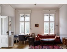 Helles Wohnzimmer mit zwei hohen Sprossenfenstern, einer roten Polstercouch, einem Holztisch und zwei schwarzen Sesseln. Zudem sind ein Holzdielentisch mit Stuhl, eine Stehlampe sowie ein Orient-Teppich und Holzboden sichtbar.