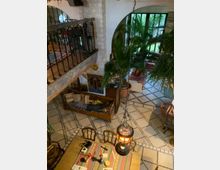 Innenansicht eines Wohnbereichs im mediterranen Stil mit gemusterten Fliesenböden, einer Holztreppe mit Geländer im oberen Bereich und einer Sitzbank mit Kissen. Große Fenster und viele Topfpalmen verleihen dem Raum eine helle und tropische Atmosphäre.