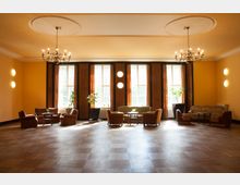 Ein geräumiger, hell beleuchteter Salon mit Holzboden, großen Fenstern und gelb gestrichenen Wänden. Der Raum ist mit mehreren Sitzgruppen aus Sesseln und Sofas sowie kleinen Tischen eingerichtet, ergänzt durch dekorative Pflanzen und hängende Kronleuchter an der Decke.