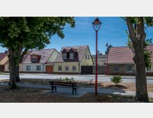 Straßenszene in einem kleinen Wohngebiet mit Einfamilienhäusern, die gelbe und beige Fassaden sowie rote Ziegeldächer haben. Im Vordergrund stehen Bäume, eine Bank und eine rote Straßenlaterne, während der asphaltierte Gehweg und die Straße die Häuser von der Sitzbank trennen.