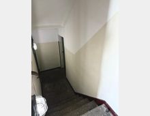 Treppenhaus mit schmaler, steinerner Treppe, die nach unten führt, flankiert von cremefarben gestrichenen Wänden und Metallgeländern. An den Wänden befinden sich Wandlampen mit schlichtem Schutzkäfig.