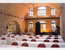 Innenansicht eines Veranstaltungsraums mit Backsteinwänden und großen Rundbogenfenstern. Es sind weiße mit Tischdecken bedeckte Tische und rote Stühle in Reihen angeordnet, während eine kleine Bühne mit Beleuchtung und Equipment im Hintergrund zu sehen ist.