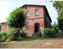 Backsteinbau mit zwei Geschossen, hohen Fenstern und einem kleinen Treppenaufgang zu einer Holztür. Das Gebäude ist von Bäumen, Büschen und Grünflächen umgeben, mit einigen Gartenmöbeln im Vordergrund.
