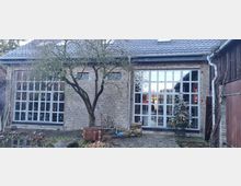 Fassade eines Gebäudes mit Backsteinmauerwerk, großen weißen Fensterelementen und einem grauen Ziegeldach. Im Vordergrund befinden sich ein kahler Baum, ein Teich und ein Weihnachtsbaum mit Lichtern.