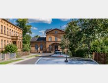 Straßenszene mit historischen Backsteingebäuden, flankiert von Grünflächen und einem Baum mit roten Beeren. Im Vordergrund sind parkende Autos und ein Gehweg zu sehen, dahinter führt die Straße auf ein größeres zweistöckiges Gebäude zu.