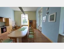 Ein heller Gemeinschaftsraum mit blau gestrichenen Wänden, großen Fenstern und hellem Holzfußboden. Im Vordergrund steht ein länglicher Holztisch mit Stühlen, im Hintergrund links eine Einbauküche und rechts ein Schrank sowie eine Musikanlage.