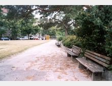 Städtischer Parkweg mit mehreren Holzbänken entlang eines gepflasterten Weges. Im Hintergrund sind Bäume, Büsche sowie eine gelbe Telefonzelle und geparkte Autos sichtbar.