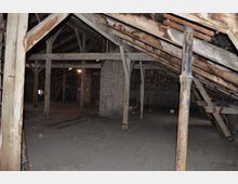 Dachboden mit sichtbaren hölzernen Trägerbalken und Stützen, zum Teil gealtert und rostig. Der Raum ist spärlich beleuchtet, hat eine Holzplanken-Bodenfläche und Wände aus unverputztem Mauerwerk.