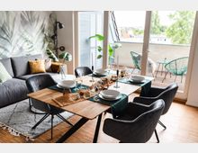 Modernes Esszimmer in einem Wohnbereich mit einem Holztisch, gedeckt für vier Personen, und schwarzen Polsterstühlen. Im Hintergrund befinden sich eine graue Couch, eine Wand mit Pflanzenmotiv-Tapete und eine große Glastür, die auf einen Balkon mit Sitzmöbeln und Pflanzen führt.