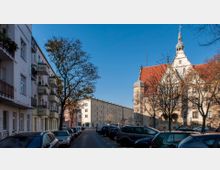 Straßenszene in einer Stadt mit geparkten Autos, links mehrstöckige Wohnhäuser mit Balkonen und rechts ein historisches Gebäude mit rotem Ziegeldach und einem Turm. In der Mitte stehen Bäume ohne Laub, und der Himmel ist klar und blau.