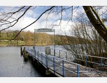 Ein Anlegersteg mit blauem Geländer führt ins Wasser, darüber ein Schild mit der Aufschrift "Anlegestelle Wirtshaus zur Fraueninsel". Im Hintergrund ist ein See mit bewaldetem Ufer und kahlen Ästen zu sehen.