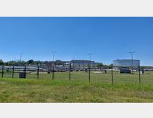 Industrieanlage mit großen, zylindrischen Tanks und einer Vielzahl von Rohren, umgeben von einem Zaun. Im Vordergrund ist eine Wiese, dahinter sind Beleuchtungsmasten, Gebäude und technische Strukturen unter klarem, blauem Himmel sichtbar.