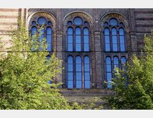 Backsteinfassade eines historischen Gebäudes mit großen, spitzbogigen Fenstern und dekorativen Mustern aus Glas. Im Vordergrund stehen grüne Bäume, die einen Teil der Fassade verdecken.