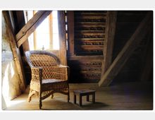 Ein Dachboden mit sichtbaren Holzbalken und Dachziegeln. Im Vordergrund stehen ein geflochtener Rattanstuhl und ein kleiner Holzhocker neben einem Fenster, durch das Tageslicht einfällt.