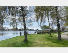 Grüne Parklandschaft mit Birkenbäumen im Vordergrund, einem Weg rechts und einem ruhigen See mit Uferbebauung und Booten im Hintergrund. Links steht eine kleine Backsteinstruktur, umgeben von Rasen und einzelnen Sitzbänken.