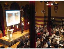 Ein Veranstaltungsraum mit dekorativen roten Vorhängen und gelben Wänden, in dem eine Person auf einer kleinen Bühne vor einem projizierten Bildschirm spricht, umgeben von einem sitzenden Publikum. Der Raum weist hohe Fenster und eine warme Beleuchtung auf.