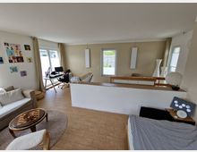 Modernes Wohnzimmer mit hellen Wänden und Holzboden, eingerichtet mit einem Sofa, Couchtisch, Schreibtisch mit Computer sowie dekorativen Elementen wie Bildern an der Wand und Stehlampen. Große Fenster lassen viel Tageslicht herein, und ein abgetrennter Bereich im Hintergrund verfügt über weitere Möbel und Dekorationen.