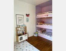 Kinderzimmer mit einem Etagenbett in lila Farbe, dekoriert mit Kissen und Decken mit Streifenmuster. Links steht eine Spielküche aus Holz vor einer weißen Wand, die mit einem Schmetterlings-Dekor und kindlicher Kunst geschmückt ist. Der Raum hat einen Holzboden und eine gepunktete Spielmatte.