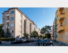 Straßenszene in einem Wohngebiet mit mehrstöckigen, farbigen Wohngebäuden; links ein beiges Gebäude mit Balkon, rechts ein gelbes Gebäude mit grünen und roten Häusern im Hintergrund. Geparkte Autos säumen die Straße, und Bäume stehen am Straßenrand.